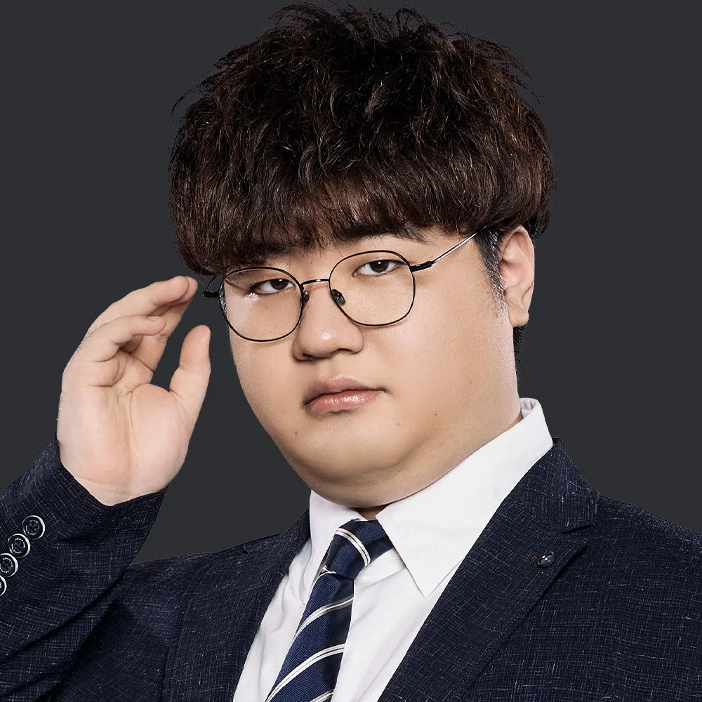 暗い背景に、スーツとネクタイを着用し丸い眼鏡をかけたアジア系の男性が写っています。右手を眼鏡に添え、短くカールした髪型が特徴的な真面目な表情のポートレートです。