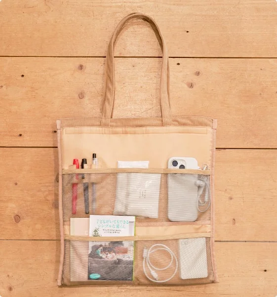 木製の背景に置かれたベージュ色のオーガナイザーバッグです。メッシュポケットにはペン、スマホ、本、充電ケーブルなどが整理されて収納されており、日常使いに便利な機能的なデザインが特徴です。