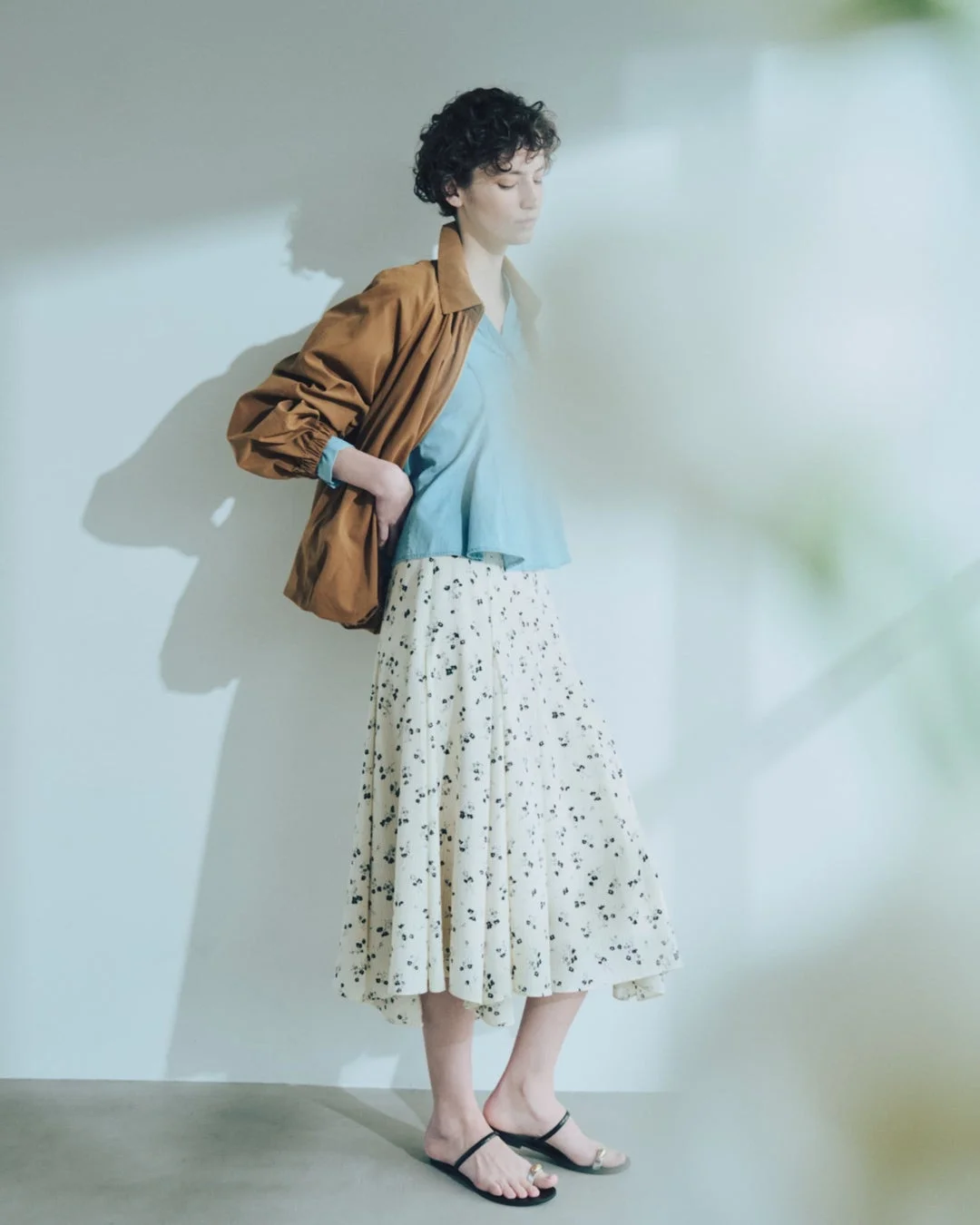 茶色のジャケットと水色のブラウス、白地に黒い花柄のロングスカートを着用した女性。