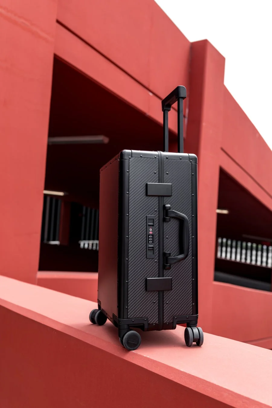 赤い幾何学的な建築物を背景に、黒いハードシェルタイプのスーツケースが置かれている写真です
