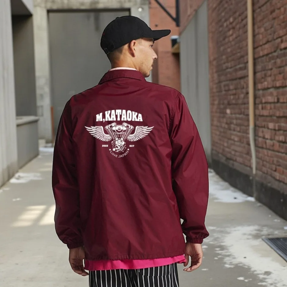 路地裏を歩く男性の後ろ姿。ボルドー色のコーチジャケットには「M. KATAOKA」の文字と翼のあるエンジン、ドクロのグラフィックがプリントされており、ストリート感のある着こなしです。