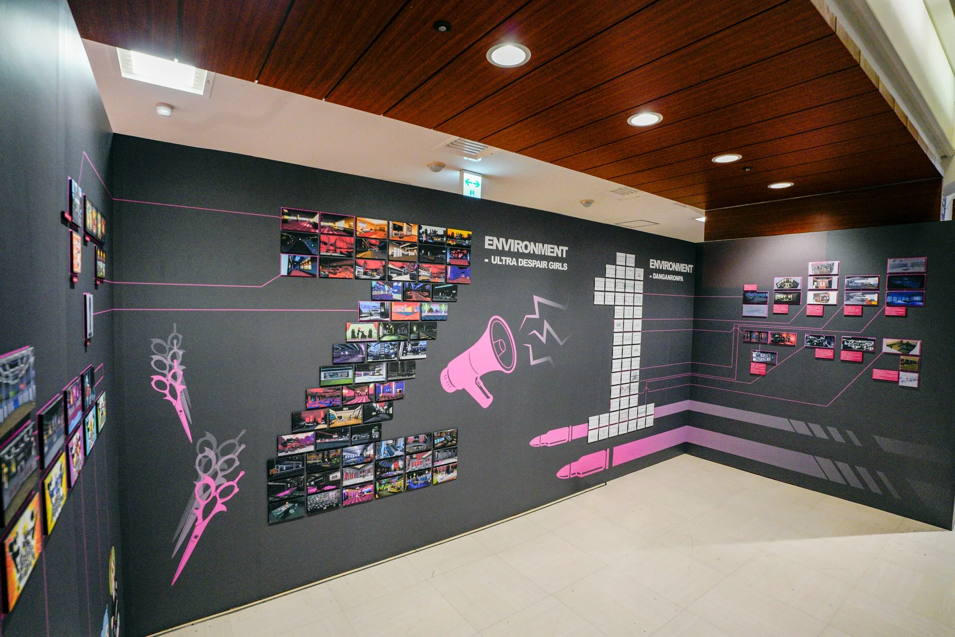 人気ゲーム「ダンガンロンパ」シリーズの環境アートを展示する空間です。黒い壁にゲームのスクリーンショットが多数飾られ、ピンクのハサミやメガホンなどのグラフィックがアクセントになっています。