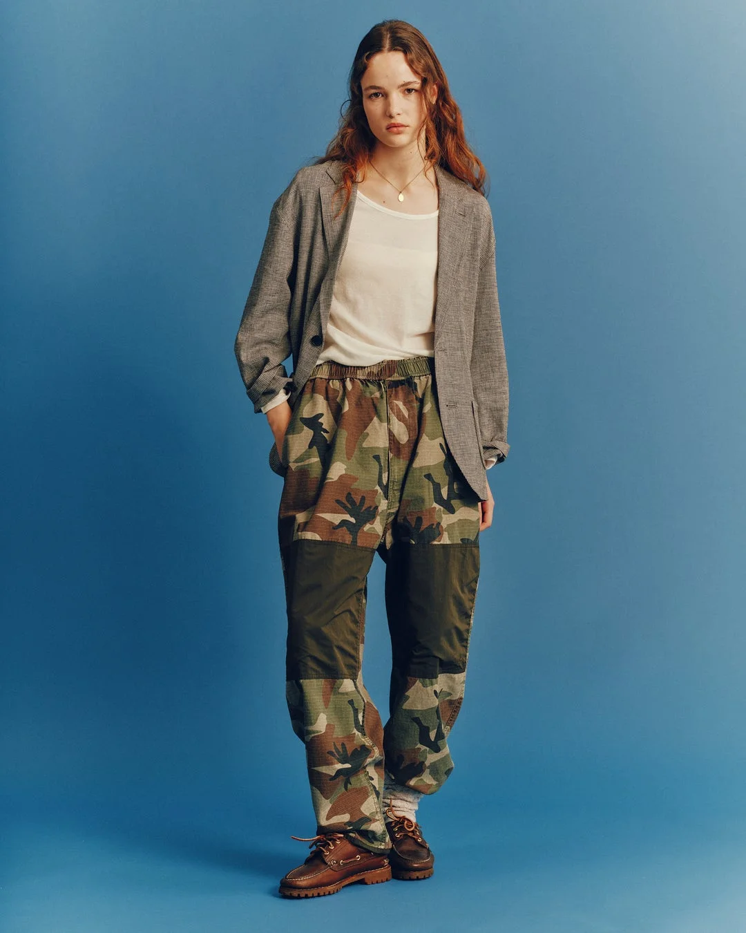青い背景の前でポーズをとる女性の全身像。彼女はカモフラージュ柄のパンツ、白いTシャツ、グレーのブレザー、そしてブラウンのボートシューズを着用しており、カジュアルながらもスタイリッシュなファッションを披露しています。