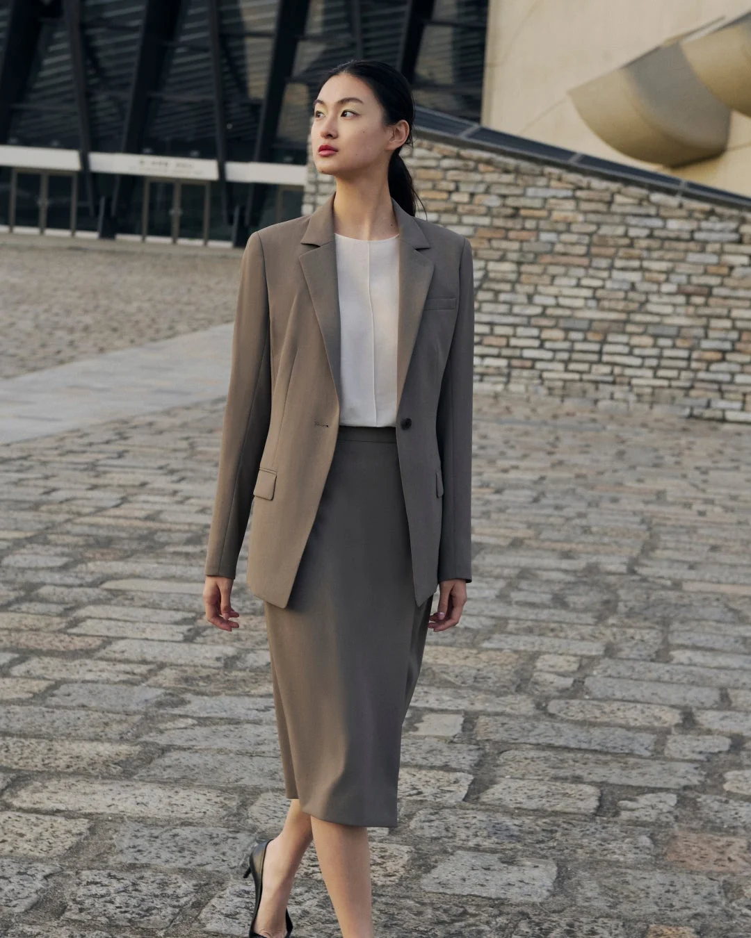 茶色のビジネススーツを着た女性が石畳の上を歩いています。彼女は自信に満ちた表情で遠くを見つめており、背景にはモダンな建物が見えます。プロフェッショナルで洗練された印象の画像です。