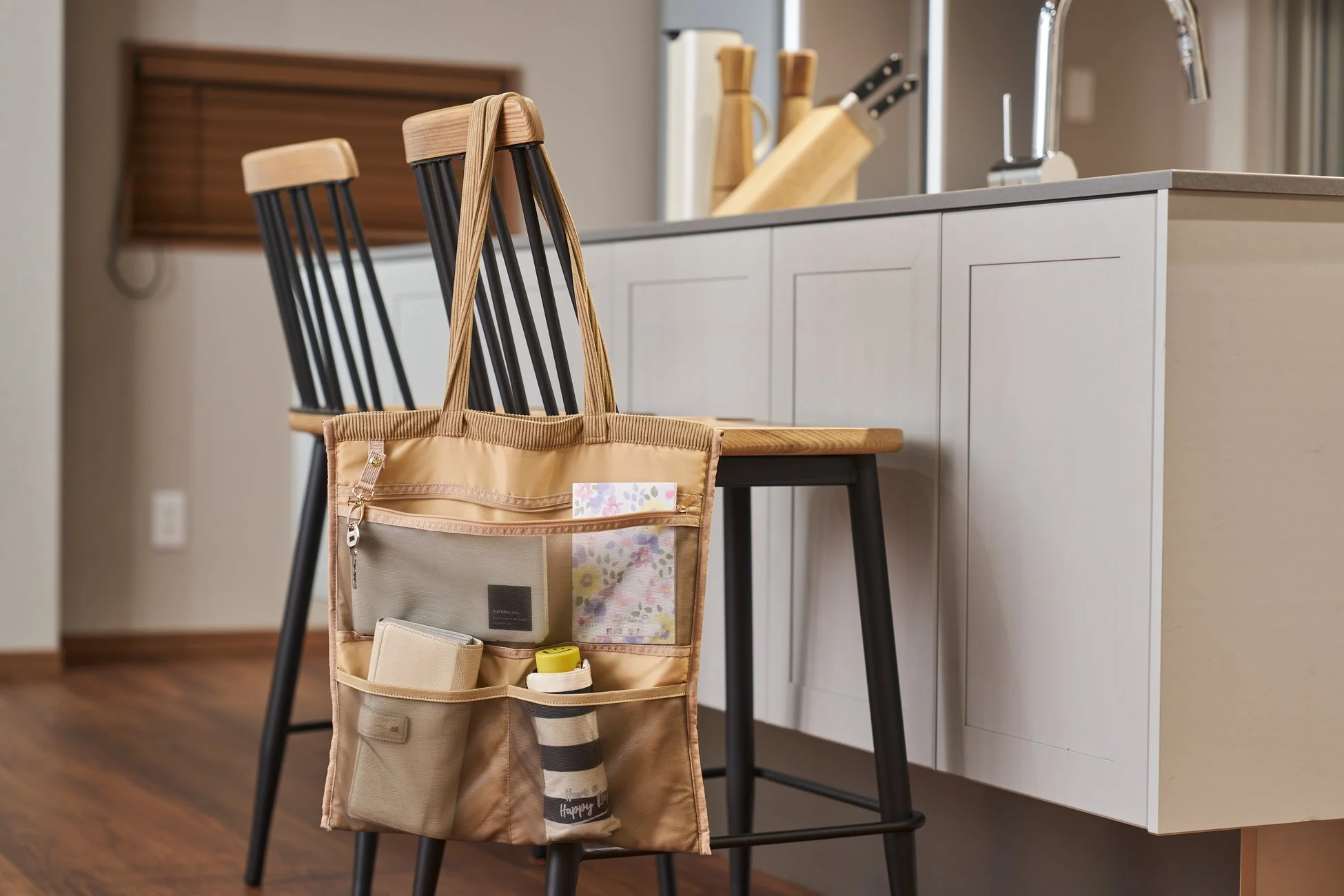 ベージュ色の多機能バッグがキッチンカウンター近くの椅子に掛けられ、財布や鍵、水筒などが整理されて収納されている様子。