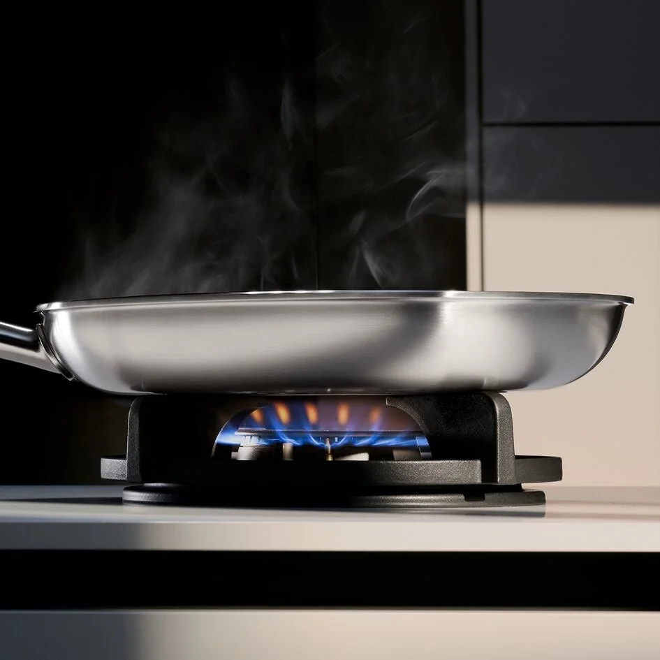ガスコンロの上でステンレス製のフライパンが加熱され、青い炎と湯気が立ち上っている様子を捉えた画像です。