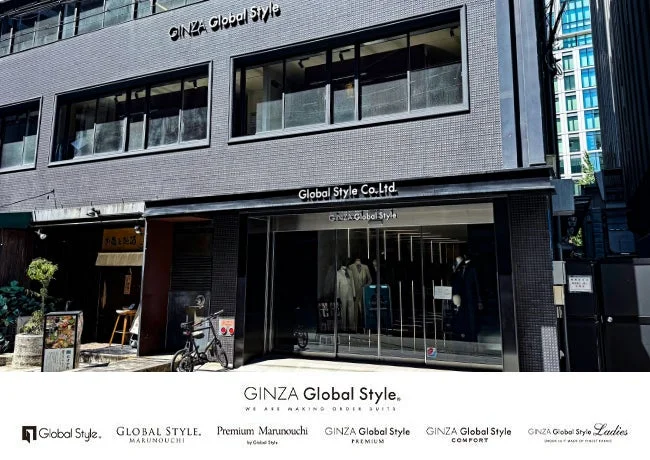 「GINZA Global Style」というオーダースーツ店の外観を捉えた画像です。モダンなグレーの建物に大きなガラス窓があり、店内にはスーツがディスプレイされています。隣には居酒屋のような店舗も見え、都市の街並みが伺えます。