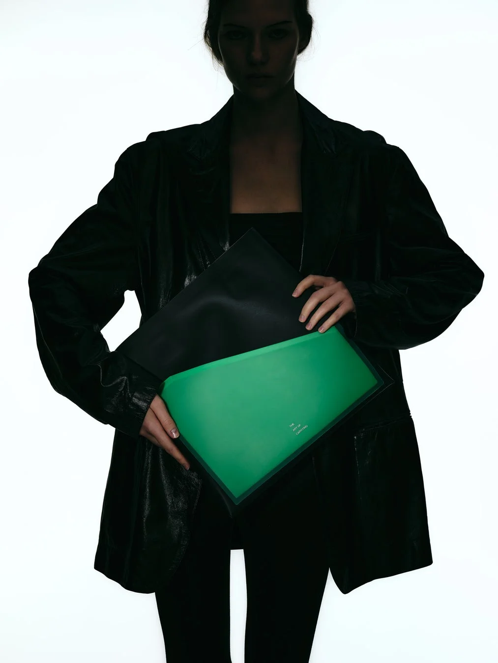 黒いジャケットを着た人物が緑色のクラッチバッグを両手で持っている画像