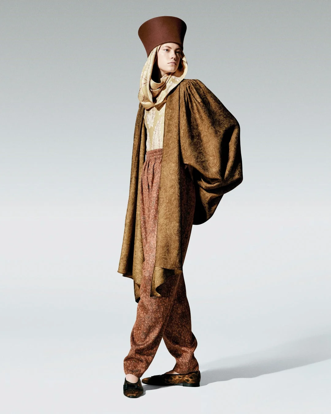 独特なブラウンの帽子とペイズリー柄のケープ、パンツを着用したモデル