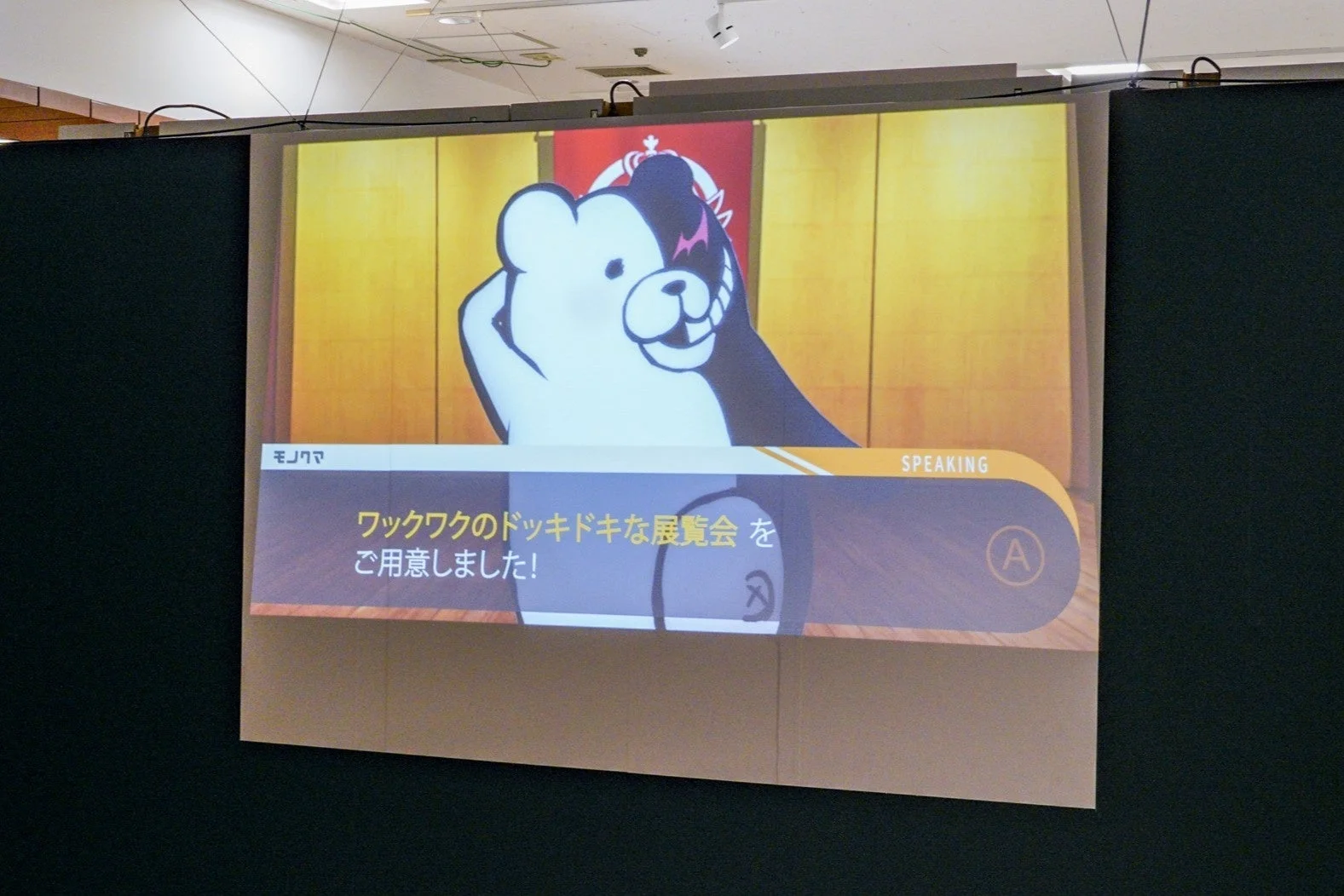 ダンガンロンパのモノクマが映し出されたプロジェクションスクリーン。ゲーム画面のような表示で、「ワクワクのドキドキな展覧会をご用意しました!」というセリフが表示されている。イベントや展示会でのゲームコンテンツ上映の様子。