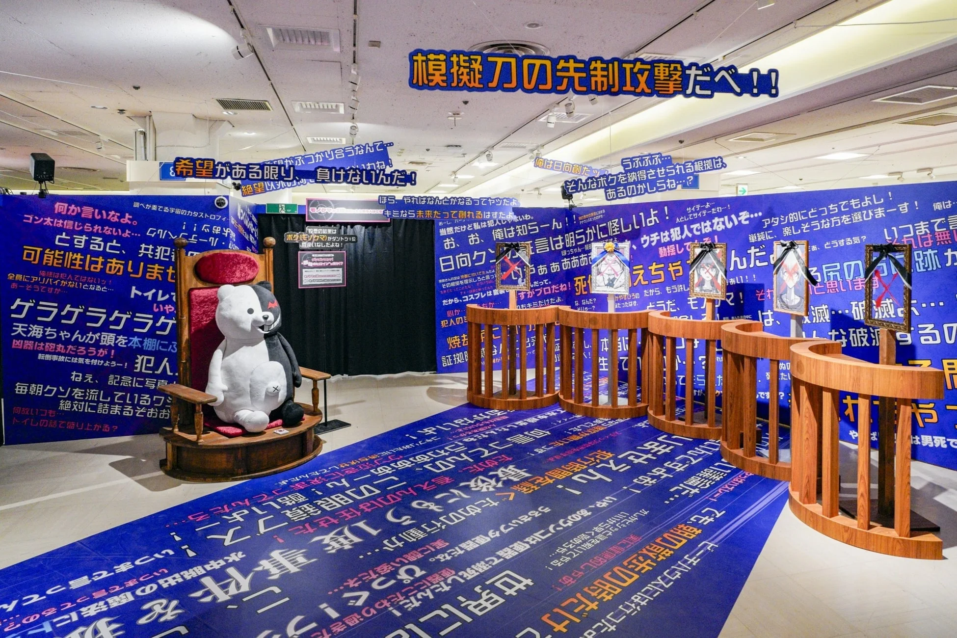 人気ゲーム「ダンガンロンパ」をテーマにした展示イベントの様子です。青い壁と床には、希望、絶望、真実、嘘、犯人といったシリーズのキーワードやキャラクターのセリフが多数書かれています。モノクマの大きなぬいぐるみと、額縁に入ったキャラクターイラストが配され、ゲーム内の学級裁判の雰囲気を再現しています。