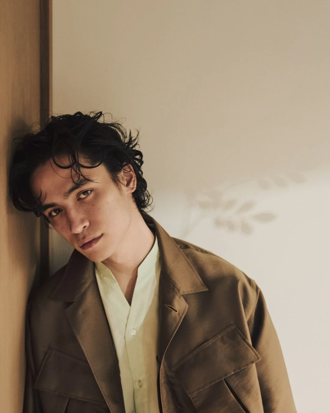 茶色のジャケットと薄緑色のシャツを着て壁に寄りかかる男性。