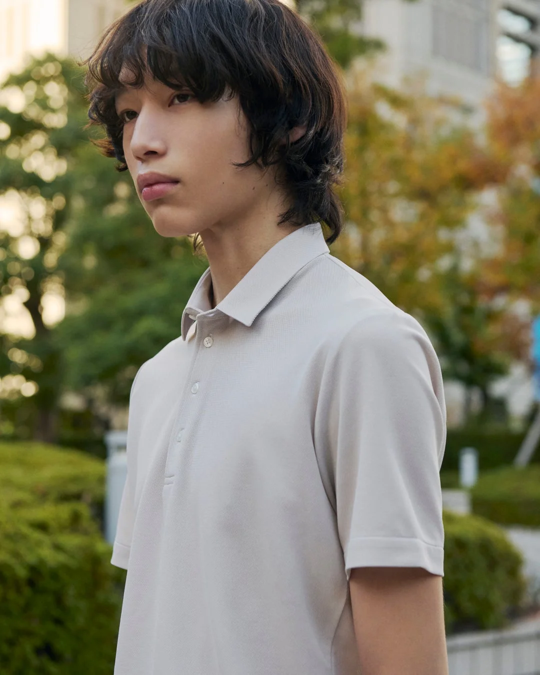 ライトグレーのポロシャツを着た若い男性が、屋外で横向きに立つポートレート。自然なウェーブヘアが特徴的で、背景には緑豊かな木々がぼかして写っている。
