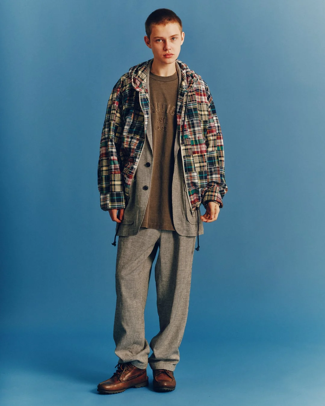 青い背景の前に立つ若い男性モデル。チェック柄のパーカーを羽織り、ブラウンのTシャツとグレーのセットアップを着用しています。カジュアルながらも洗練された秋冬のストリートファッションを表現しています。