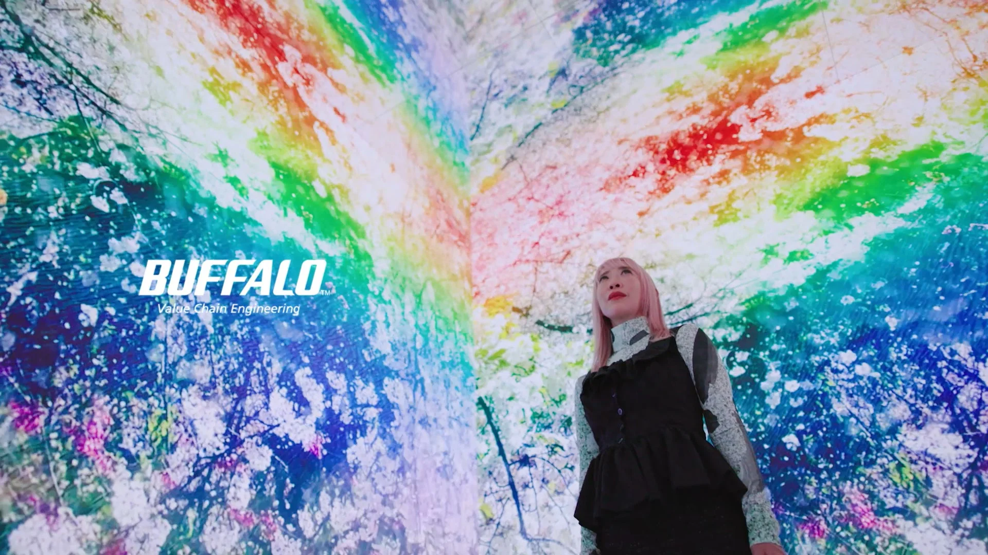 桜のような模様と虹色のグラデーションが広がる没入型デジタルアート空間を見上げるピンク髪の女性