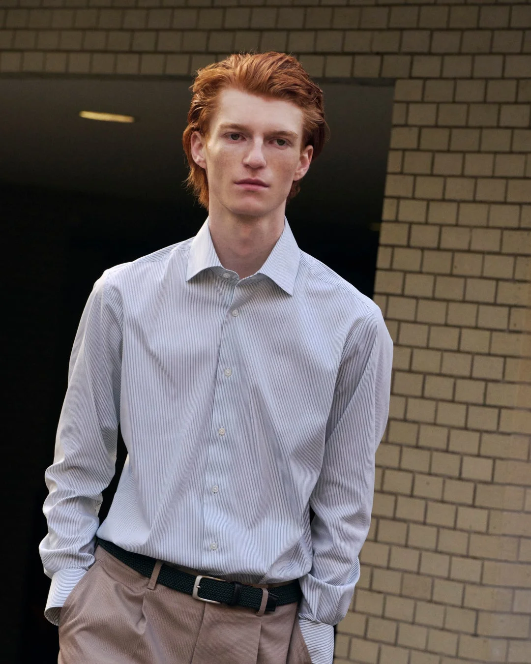 赤毛の男性がストライプ柄のシャツとベージュのパンツを着用し、屋外でカメラを見つめているポートレートです。都会的な背景が特徴的で、洗練されたカジュアルスタイルを表現しています。