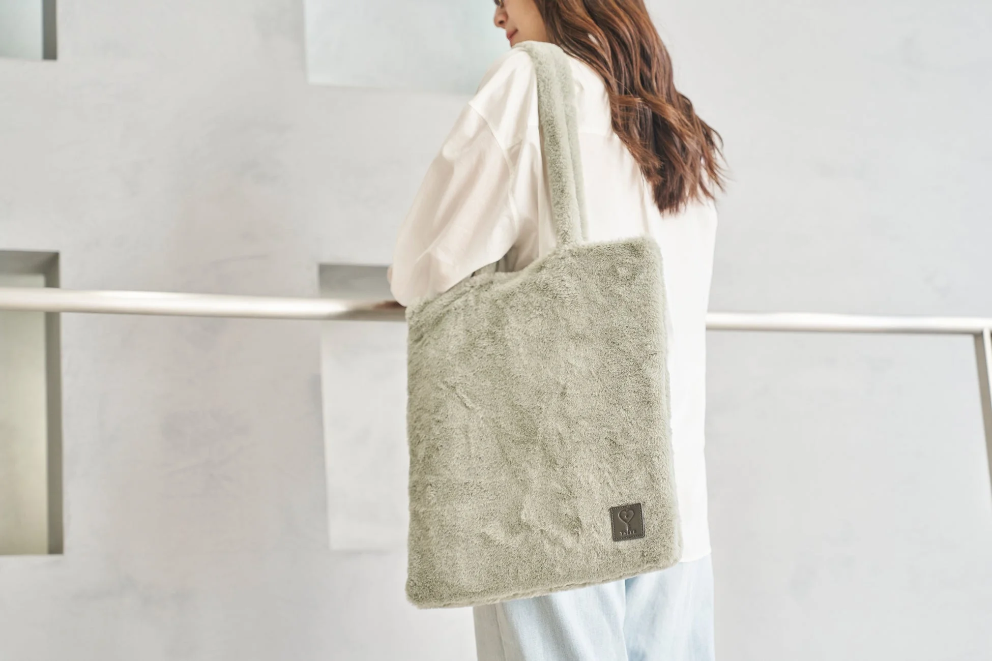 白いシャツと淡い色のパンツを着用した女性が、ライトグリーンのふわふわしたトートバッグを肩に掛けています。カジュアルで暖かみのある冬のファッションスタイルです。