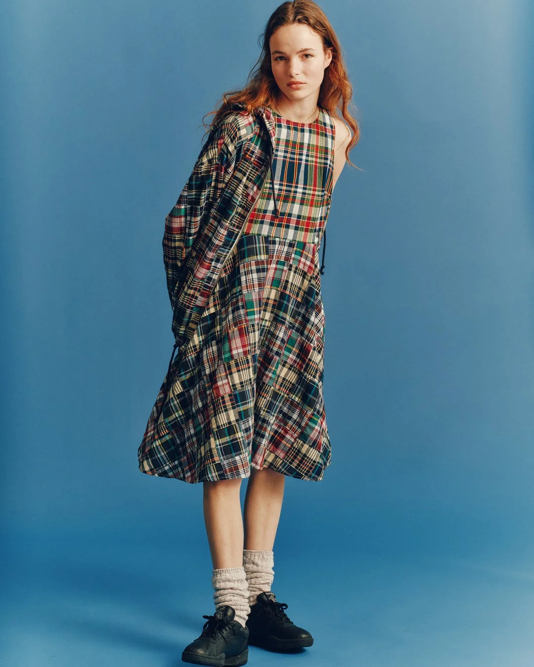 青い背景のスタジオで、チェック柄のワンピースとジャケットを着用し、スニーカーとソックスを合わせたカジュアルな服装の女性が立っています。彼女はカメラをまっすぐ見ています。