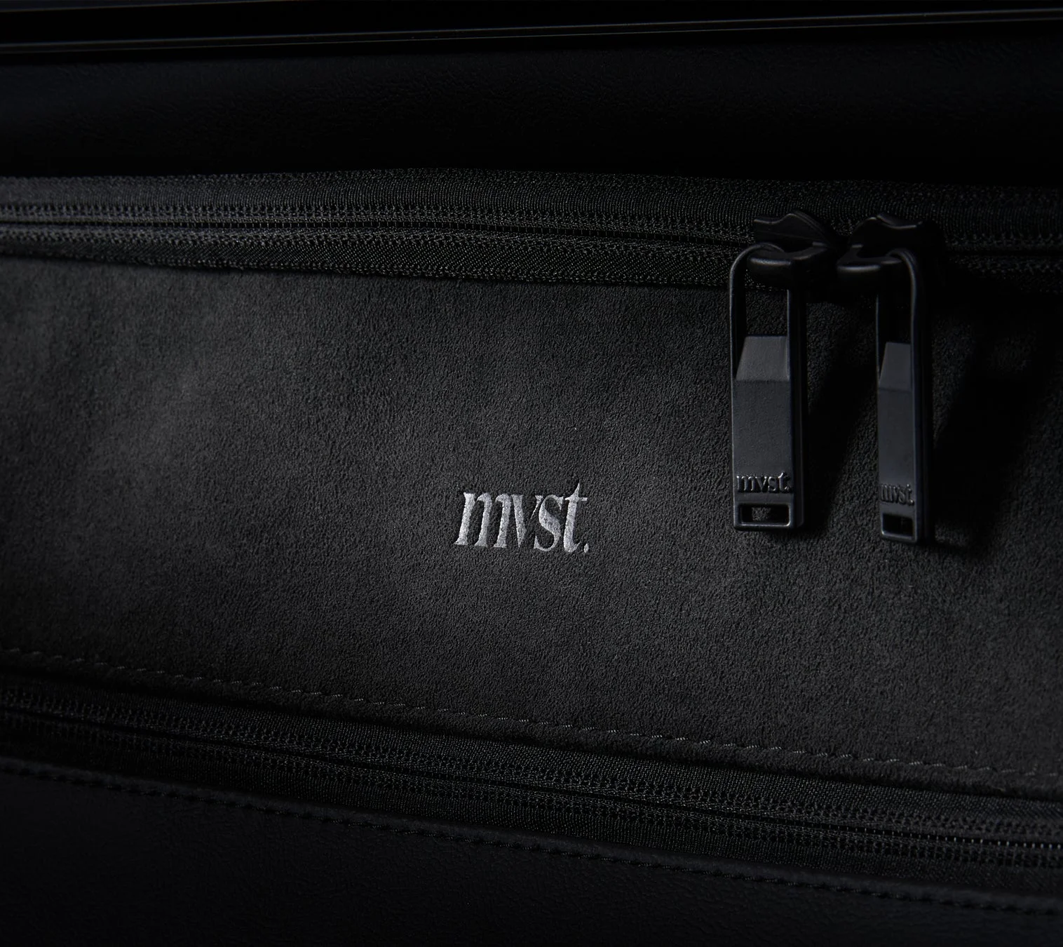 黒い質感のある素材に「mvst.」のロゴがエンボス加工され、ジッパーの引き手にも同じロゴが刻印されているクローズアップ画像です