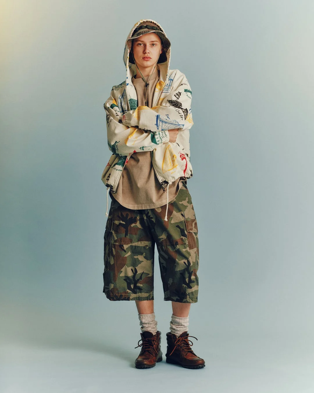 水色の背景の前でカジュアルな服装をした若者が腕を組んで立っている全身ポートレート。柄物のフーディー、迷彩柄のショートパンツ、茶色のブーツを着用しています。