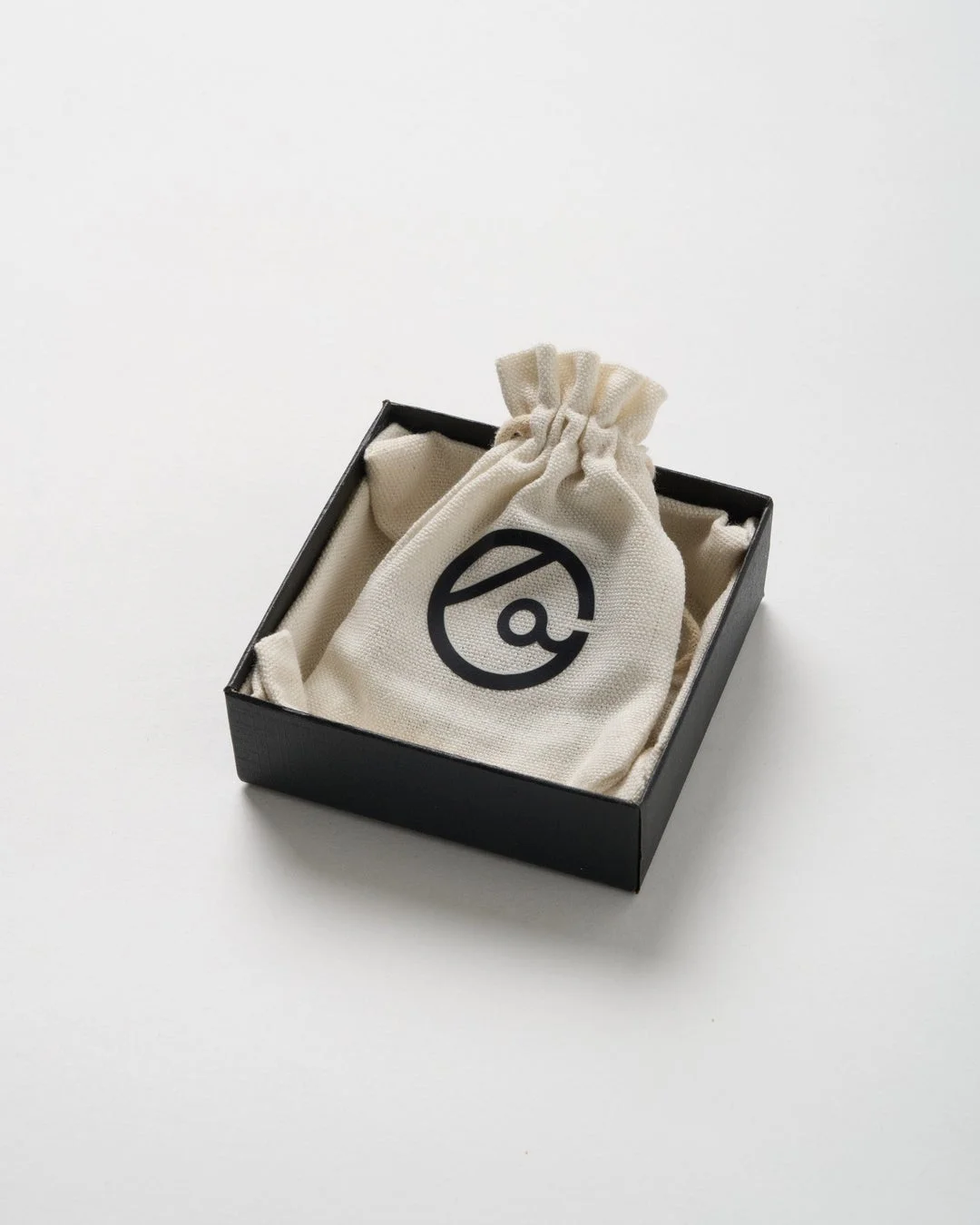 黒い箱の中にベージュの巾着袋が入っており、袋には黒い抽象的なロゴマークが印刷