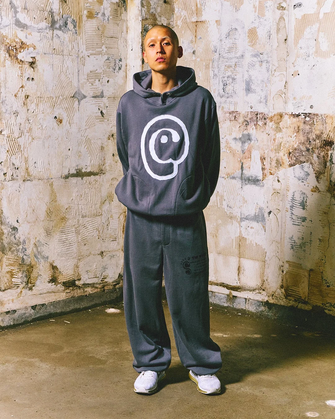 グレーのフーディーとスウェットパンツを着用した男性が、荒廃した壁の前で立っている全身像です