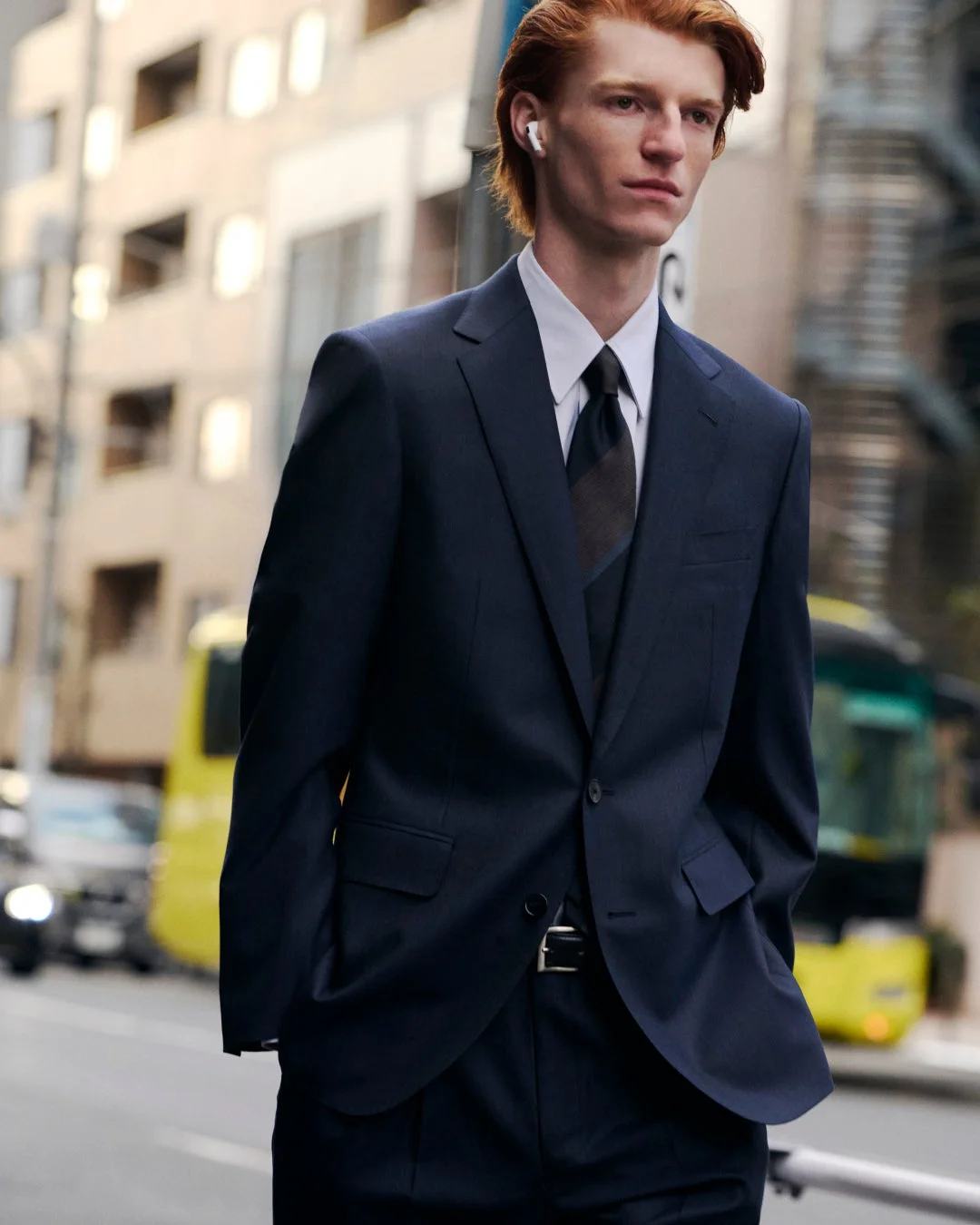 紺色のスーツを着た赤毛の男性が、街中で白いイヤホンを装着して立っている。背景には建物や黄色いバスが見え、洗練されたビジネススタイルが印象的。