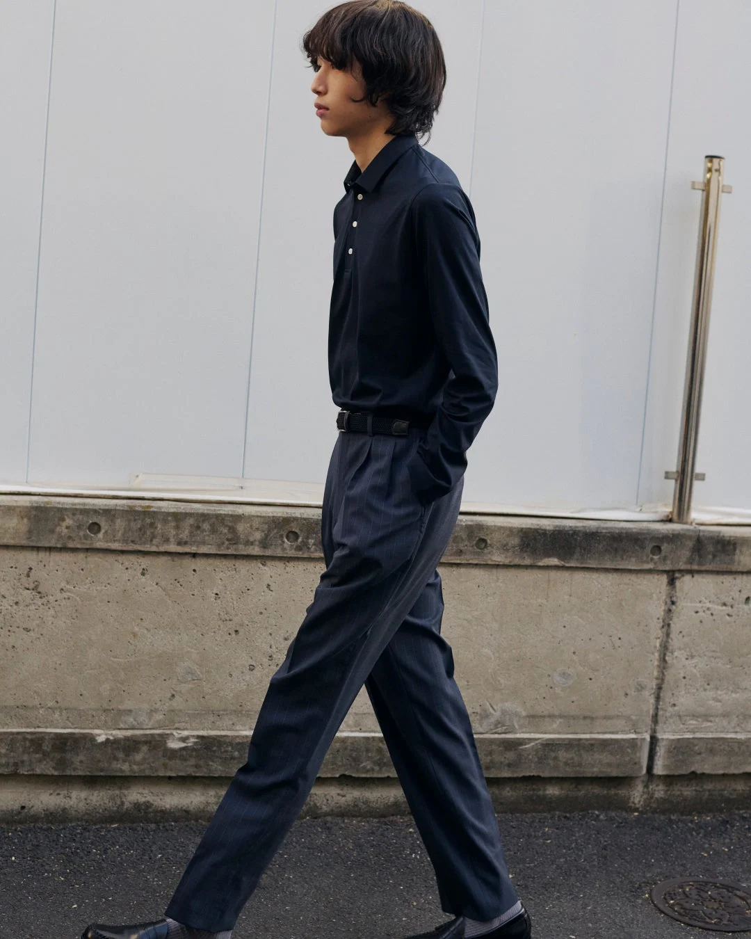紺色の長袖ポロシャツとストライプのスラックスを着用した若い男性が、街中を歩いている全身像。横顔で左を向いており、手はポケットに入れている。カジュアルながらも落ち着いた雰囲気です。