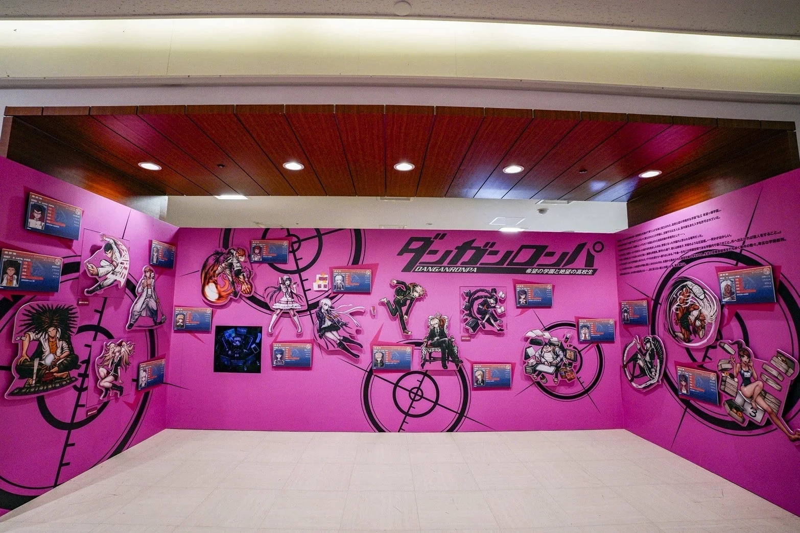 人気ゲーム「ダンガンロンパ」の展示スペースを捉えた画像です。鮮やかなピンクの壁に多数のキャラクターパネルとゲームロゴが飾られ、作品の世界観が表現されています。