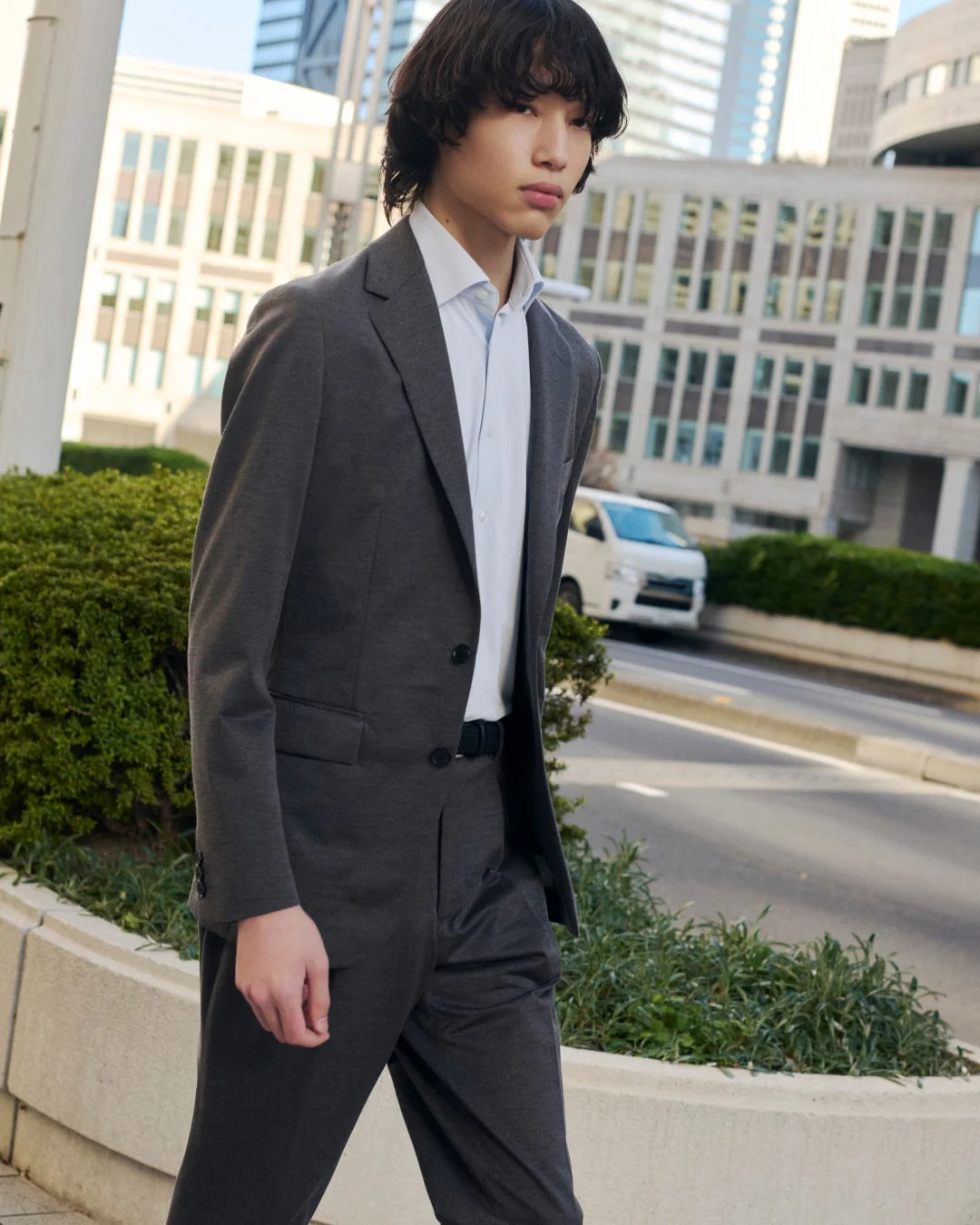 都会の街路を歩く若い男性の姿を捉えた一枚。チャコールのカジュアルなスーツに白いシャツを合わせ、洗練されたビジネススタイルを披露しています。背景には近代的なビルが立ち並び、都会的な雰囲気を強調しています。
