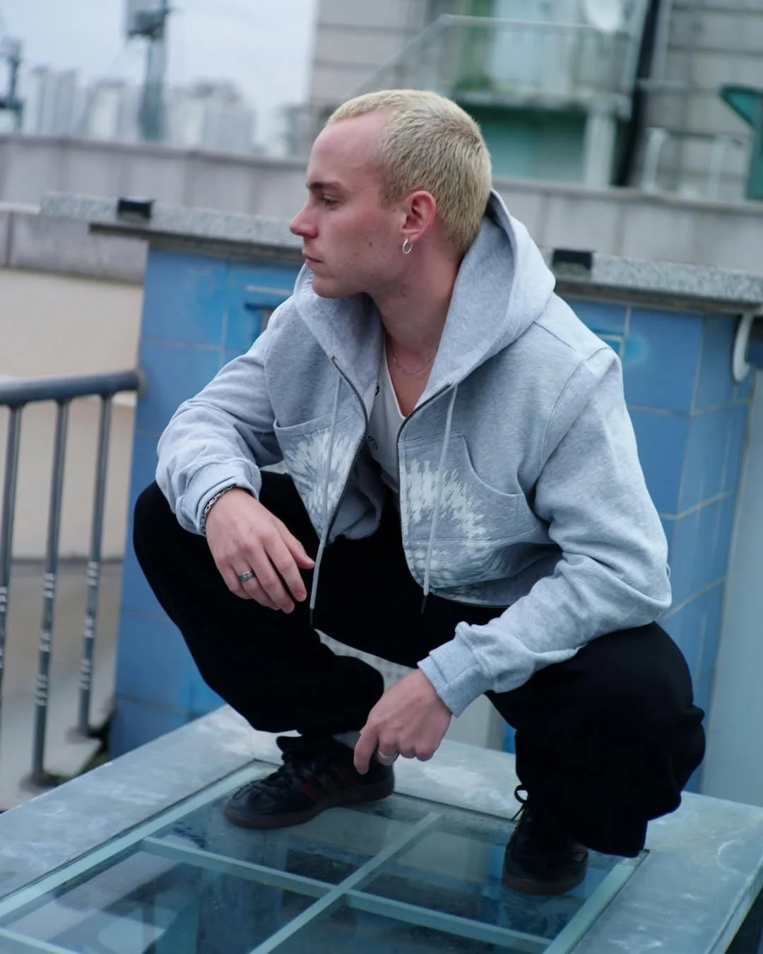 金髪の若い男性が灰色のパーカーと黒いパンツを着用し、屋上のガラス張りの床にしゃがみ込んでいる様子