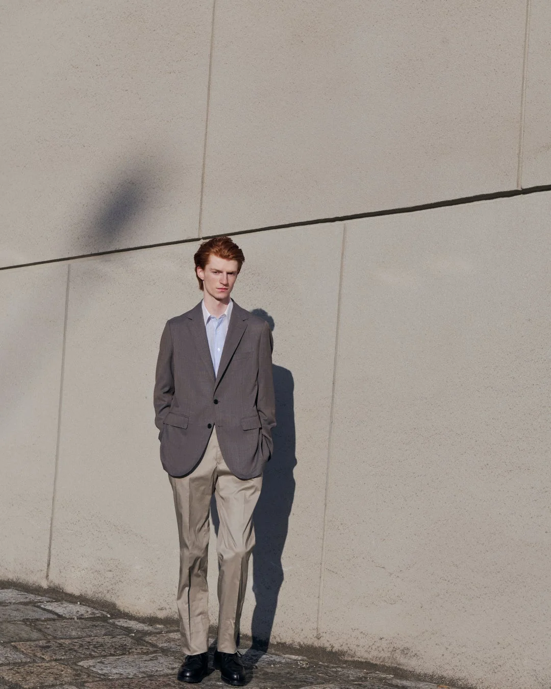 グレーのジャケットとベージュのパンツを着た赤毛の男性が、コンクリートの壁を背景に立っている。日差しが強く、壁には男性の影が映し出されている。落ち着いた雰囲気のファッションポートレート。