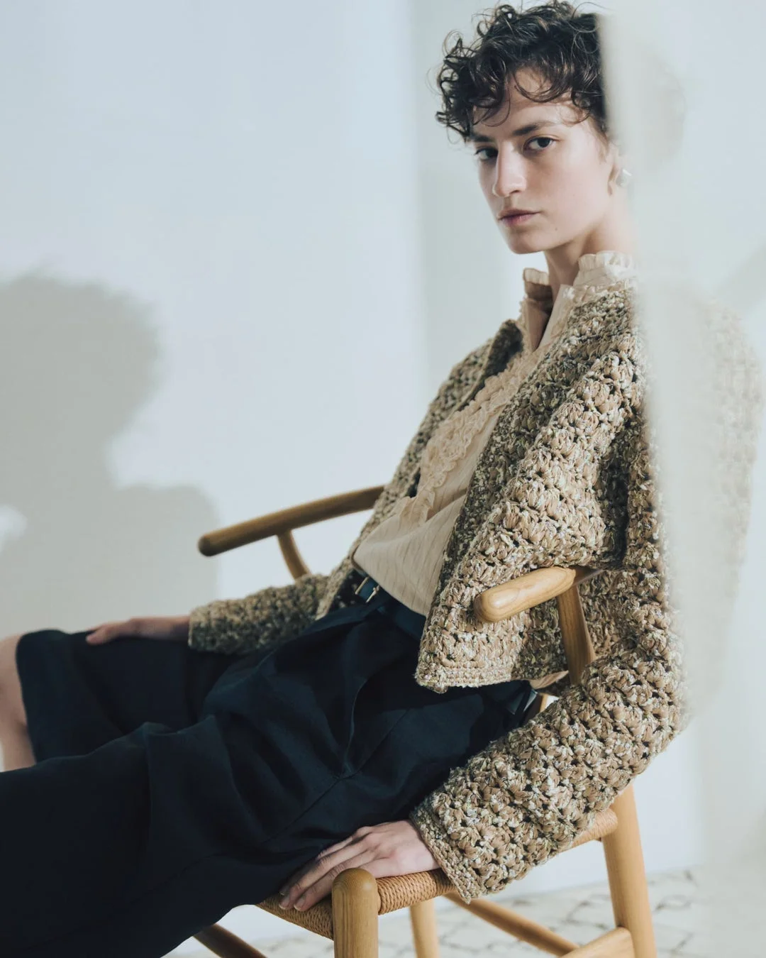 ベージュのジャケットとダークカラーのパンツを着用し、木製の椅子に座るショートヘアの女性。