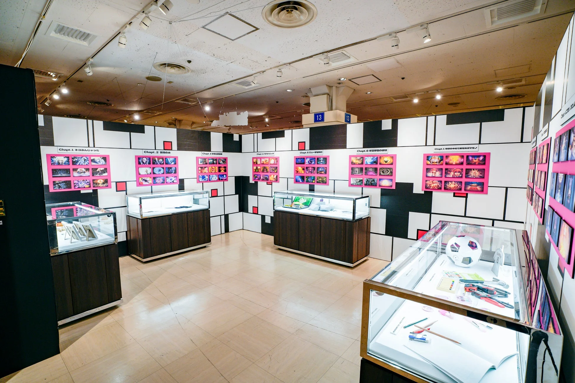 モンドリアン風の白黒赤のグリッド模様の壁面が特徴的な展示会場です。複数のガラスケースが並び、壁にはピンクの額縁に収められたカラフルなイラストやアニメーションの場面が日本語の章タイトルと共に展示されています。現代アートやイラストレーションの展覧会と思われます。