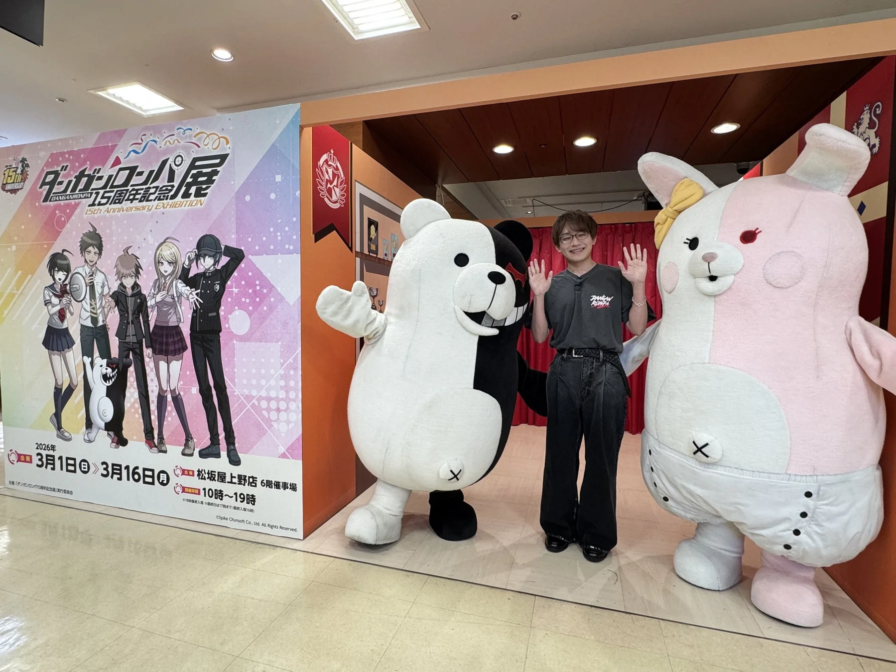 ダンガンロンパ15周年記念展で、来場者がモノクマとモノミの着ぐるみと共にポーズをとっています。背景にはイベントのロゴとキャラクターが描かれた大型パネルがあり、開催情報も確認できます。