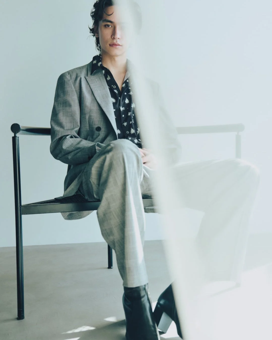 グレーのスーツと柄物の黒シャツを着て椅子に座る巻き髪の男性。