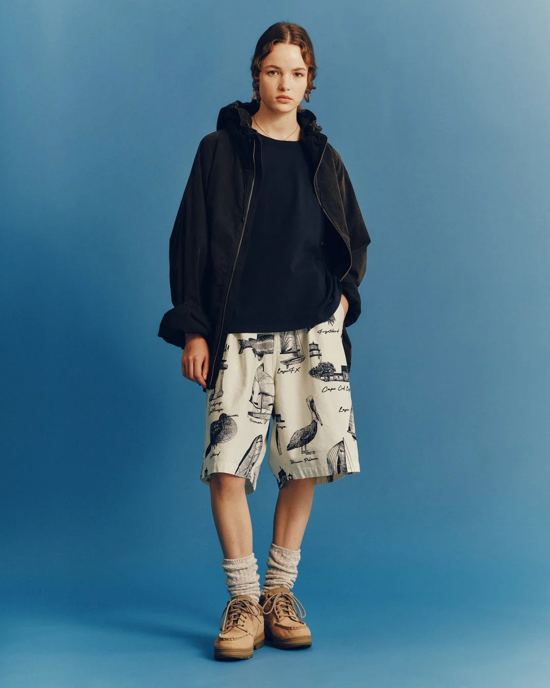 青い背景の前で、黒いジャケットとTシャツ、そして船や鳥、灯台などのイラストが描かれたクリーム色のショートパンツを着用した若い人物のポートレート。カジュアルでマリンテイストな夏のファッションスタイルです。