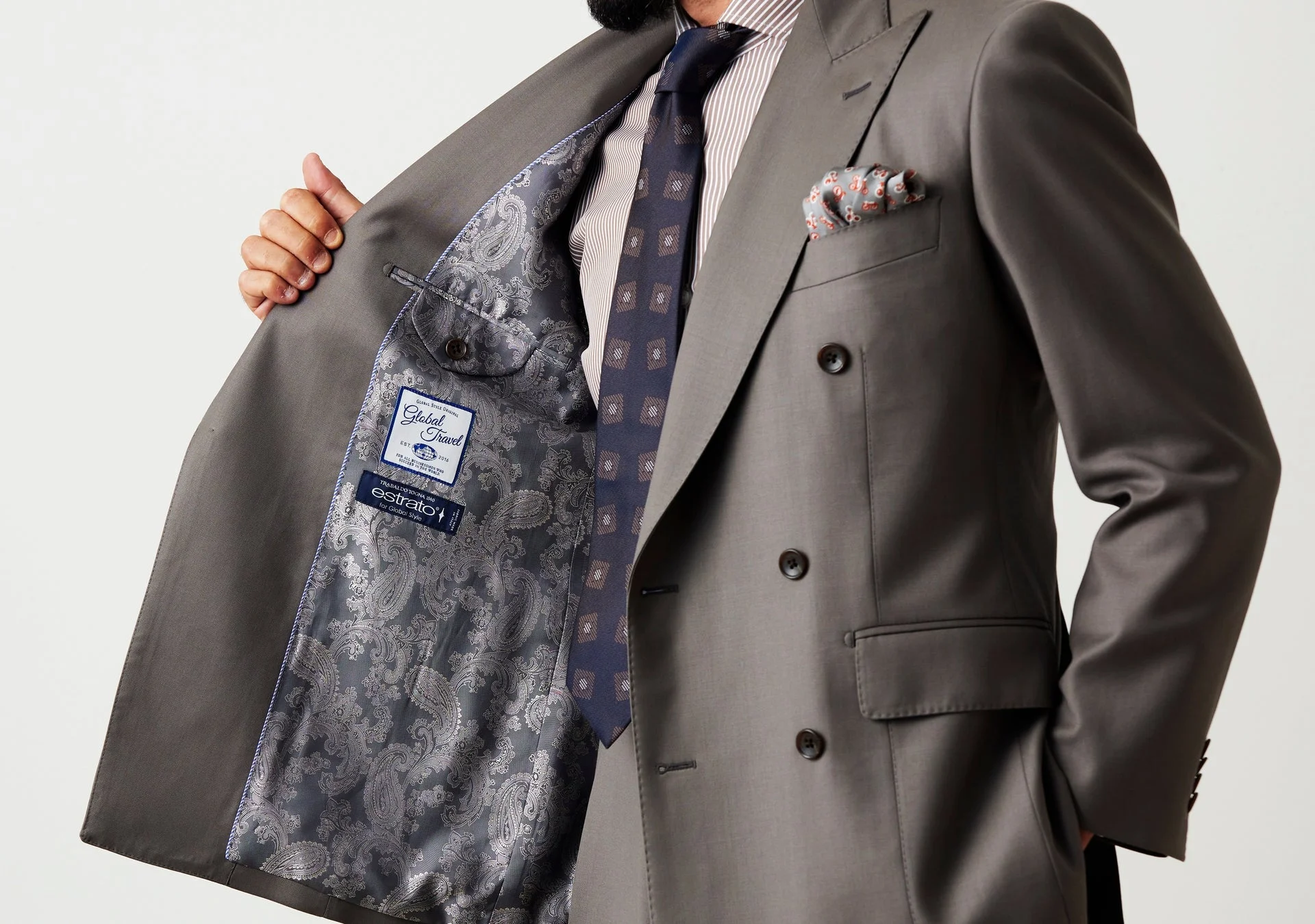 男性がグレーのダブルブレストスーツを着用し、裏地のペイズリー柄やブランドタグが見えるようにジャケットを広げている様子。ビジネスシーンに適した上品な装い。