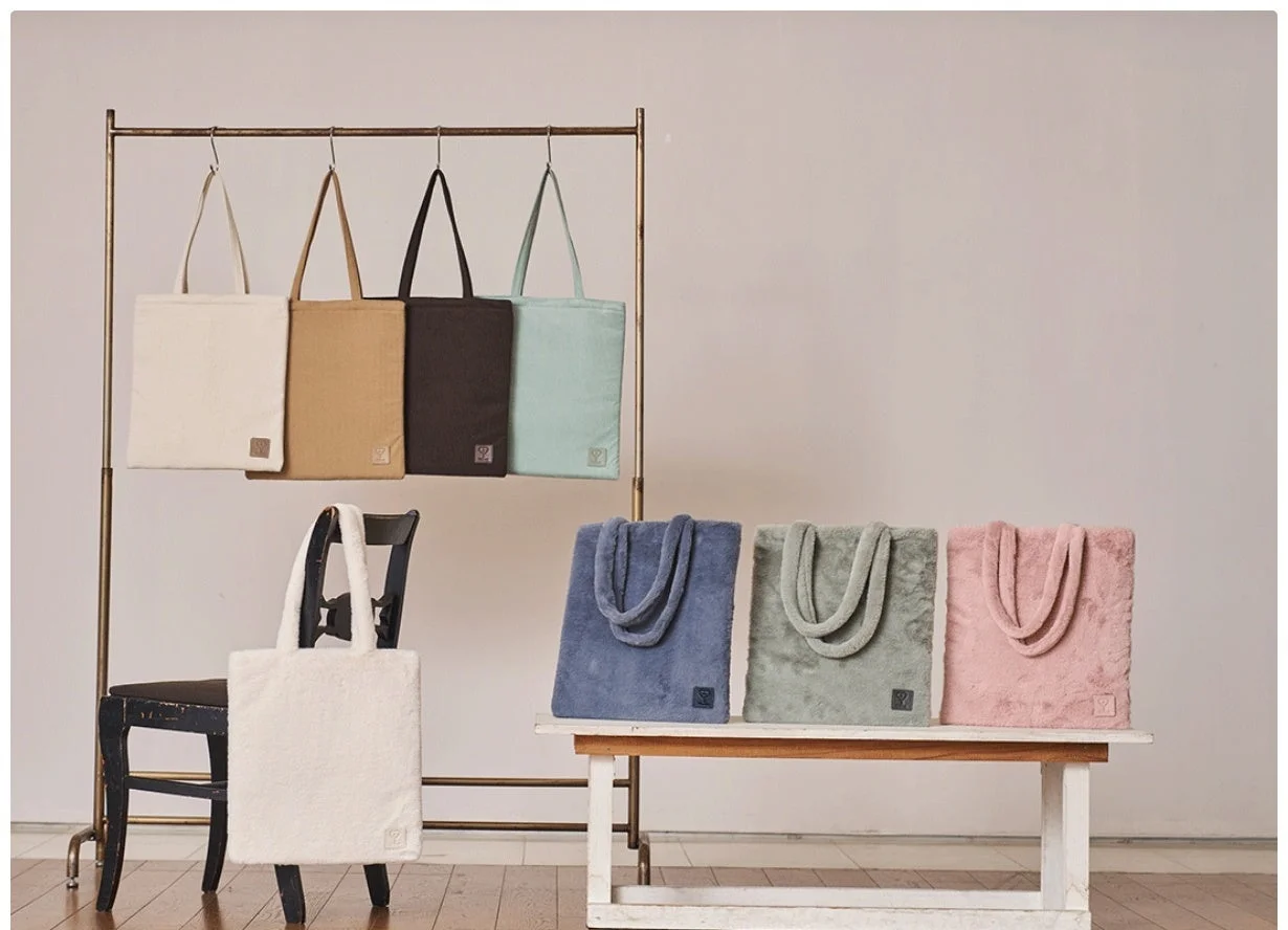 室内で様々な色のトートバッグが展示されている様子です。ハンガーラックにはシンプルな布製バッグが4つ、椅子とベンチにはふわふわした素材のバッグが5つ並べられており、商品の魅力を伝えています。
