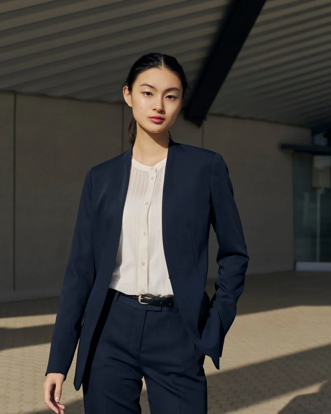 紺のノーカラージャケットとパンツのセットアップを着た女性が、屋外でカメラを見つめている。ビジネスシーンにも合うスマートな装い。
