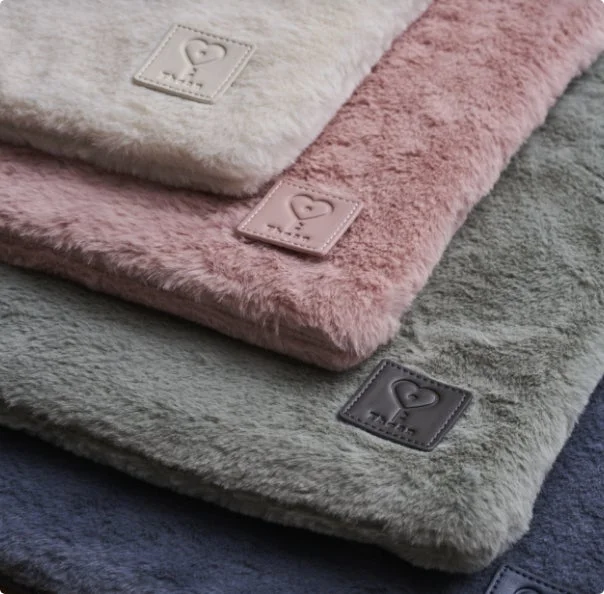 クリーム、ピンク、グレー、青系の複数の色合いの、ふわふわとした柔らかな素材の布製品が積み重ねられている様子。