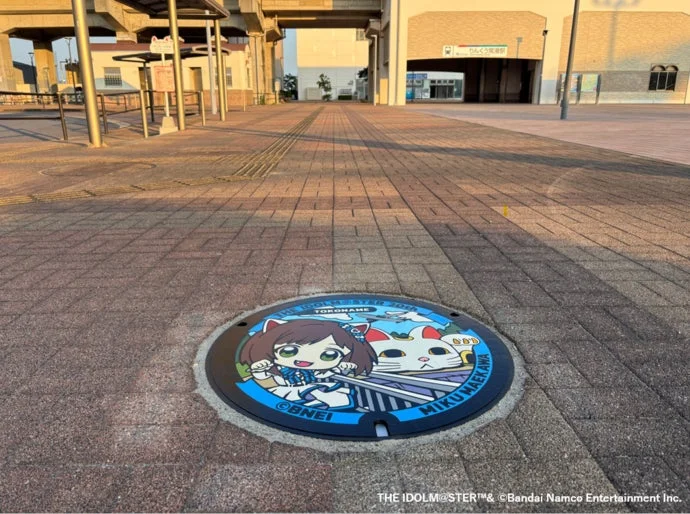 夕暮れ時のりんくう常滑駅前の広場に設置された、「アイドルマスター シンデレラガールズ」の前川みくと猫が描かれたカラフルなデザインマンホール