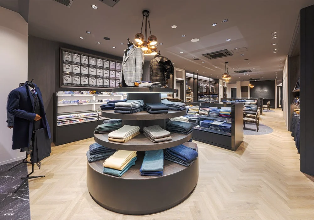 モダンで上質なメンズアパレル店舗の内部風景です。スーツ、コート、シャツ、ネクタイなどが美しく陳列されており、中央には折りたたまれた生地が円形什器に並べられています。洗練された雰囲気のショップです。