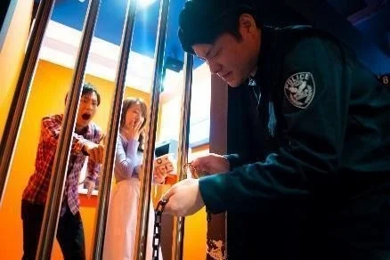 鉄格子の中にいる男女が驚いた表情を見せる中、警察官のような制服を着た男性が外から鎖を操作しています。脱出ゲームや監獄をテーマにしたアトラクションの一場面のようです。
