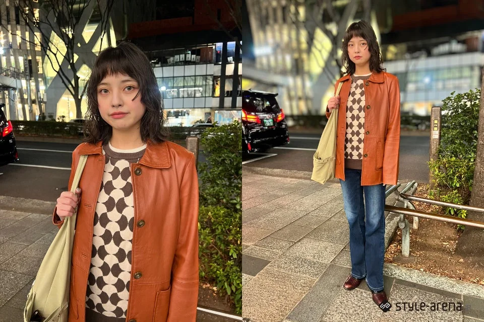 夜の街角に立つ若い女性のポートレート。ブラウンのレザージャケットと幾何学模様のニット、ブルージーンズを合わせたカジュアルながらもスタイリッシュな装いで、ベージュのトートバッグを肩にかけている。背景には都市の建物と街灯が見える。