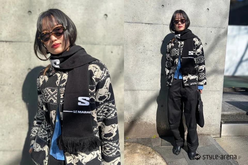 コンクリートの壁を背景に、柄物のジャケットと黒いマフラー、サングラスを身につけた女性のストリートスナップ。黒いパンツと足袋シューズを合わせた、個性的な冬のファッションコーディネートです。